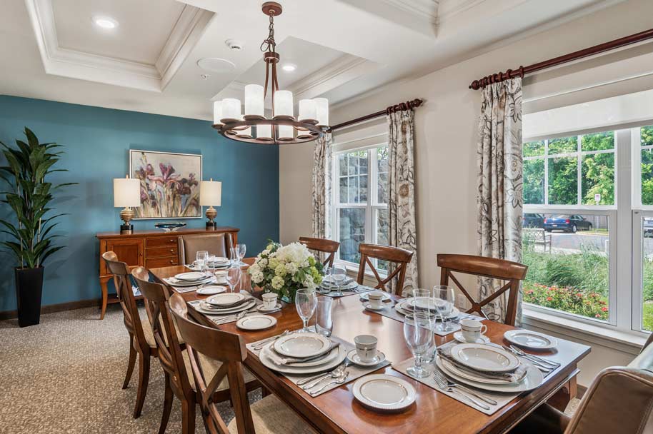 A dining room with a large wooden table set for eight, featuring elegant tableware and a floral centerpiece. The room has a blue accent wall with framed artwork, a sideboard with lamps, large windows with patterned curtains, and a ceiling light fixture.