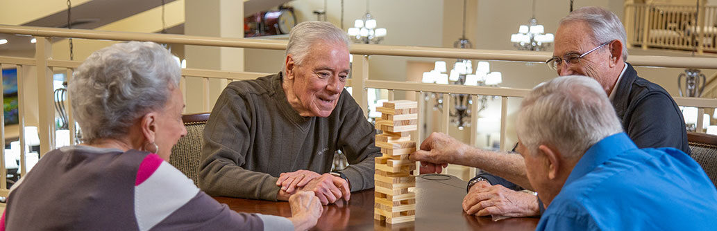 Four elderly individuals sit around a table playing a game of Jenga. They are indoors, and all appear engaged and happy. One man is reaching for a block in the tower, while the others watch with anticipation. The environment is well-lit and cozy.