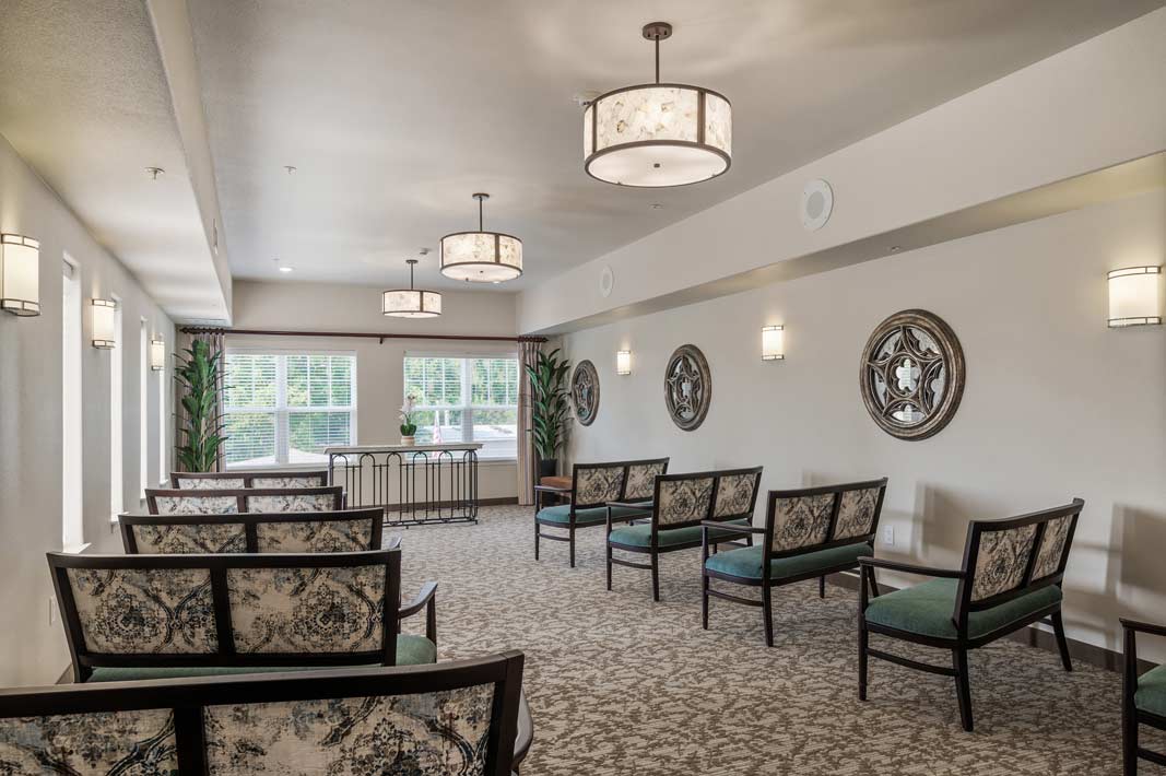 A bright and spacious room featuring rows of patterned chairs facing windows with white blinds and greenery outside. The walls are decorated with round mirrored wall art, and ceiling lights add a warm ambiance. The design is modern and inviting.