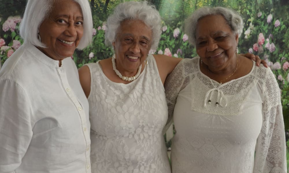 Three elderly women are standing closely together, smiling warmly. They are dressed in white, with a backdrop of a vibrant green garden and pink flowers. The women appear joyful and content as they pose for the photo.