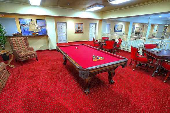 A room with red carpeting features a pool table with red felt, set up for a game. Around the room are red upholstered chairs and wooden tables. Wall art includes paintings of boats. Large windows and mirrored walls enhance the room's spacious feel.