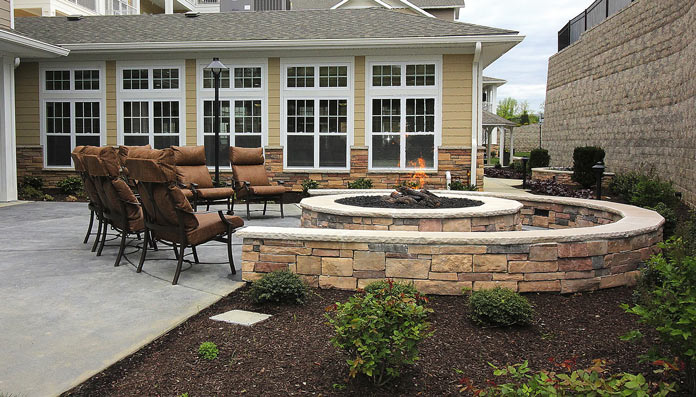 A cozy outdoor patio featuring a circular stone fire pit surrounded by brown cushioned metal chairs. The backdrop includes a building with many windows, a lamp post, and manicured garden beds with shrubs.