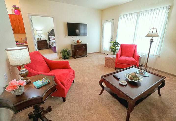 A cozy living room with beige carpet featuring two red armchairs, a wooden coffee table, and a wooden side table with a lamp and books. There's a wall-mounted TV, a dresser, a potted plant, and a doorway leading to a bedroom with a visible bed.