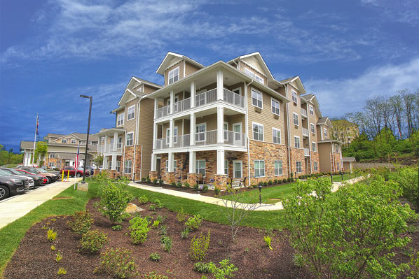 A modern, multi-story apartment building with balconies under a blue sky. The building's exterior features light-colored siding and stone accents. In the foreground, there's a landscaped area with shrubs and a parking lot with several cars to the left.