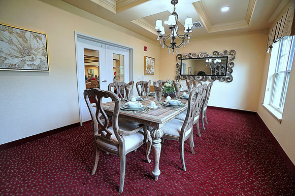 A well-lit dining room with a long, wooden table set for eight. The room features a chandelier, ornate mirror, framed artwork, and large window. Chairs have cushioned seats and the table is set with plates, bowls, and cups. The carpet is red with a subtle pattern.