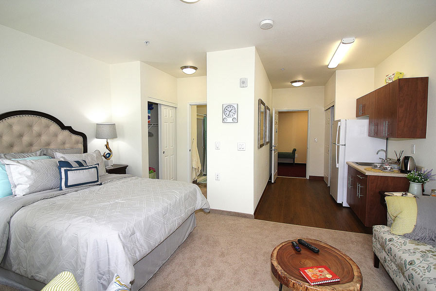 A well-lit studio apartment with a bed on the left, a small kitchen on the right, and a seating area featuring a round wooden coffee table. There's a bathroom door near the kitchen and an open closet next to the bed. The decor is modern and cozy.