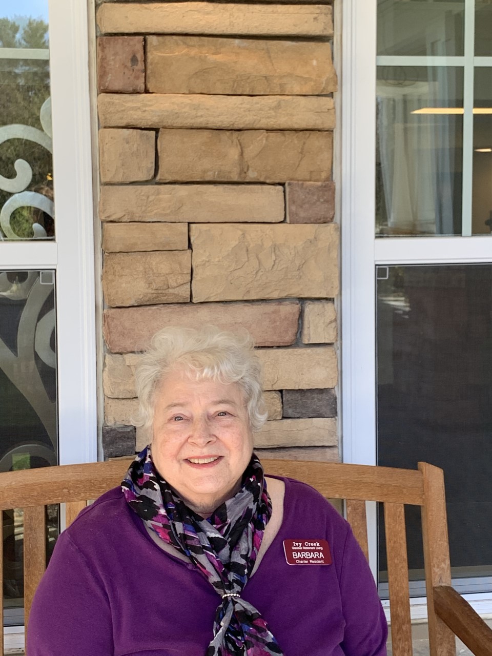 An elderly woman with white hair is sitting on a wooden bench in front of a stone wall. She is wearing a purple top and a patterned scarf, smiling at the camera. There are windows behind her with white frames and curtains visible through the glass.