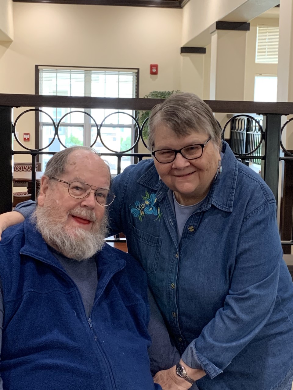 An elderly man with a white beard, wearing glasses and a blue vest, is seated next to an elderly woman with short hair, wearing glasses and a blue denim shirt with floral embroidery. The woman has her arm around the man, and they are both smiling at the camera.