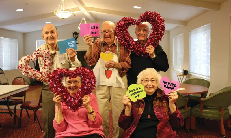 A group of cheerful elderly people holding heart-shaped decorations and colorful signs with messages, gathered in a warmly lit room. Two people are standing at the back, and three are seated at the front, all smiling and appearing to enjoy the festive atmosphere.
