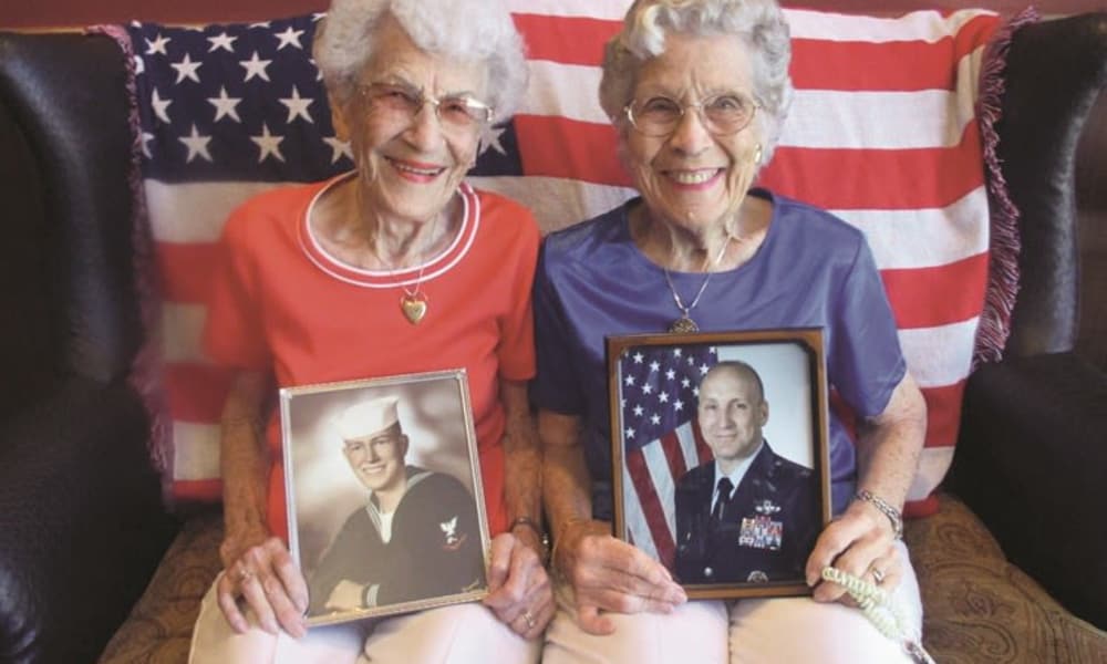 Two elderly women are sitting side by side on a couch with an American flag draped behind them. Both are smiling and holding framed photographs of men in military uniforms. One wears a red shirt, the other a blue shirt, symbolizing patriotism.