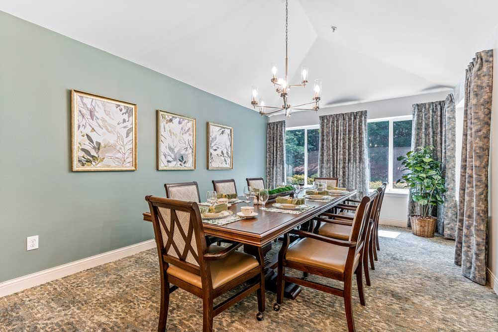 A dining room with a long wooden table set for six, featuring elegant tableware. The room has green walls adorned with three framed botanical prints, a modern chandelier, patterned curtains, and a large window with a view of the greenery outside.