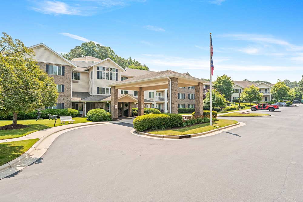 A multi-story senior living facility with a large covered entrance. Well-maintained landscaping features shrubs and trees. An American flag is near the entrance. Cars are parked along the driveway, which curves around the front of the building. Blue sky overhead.