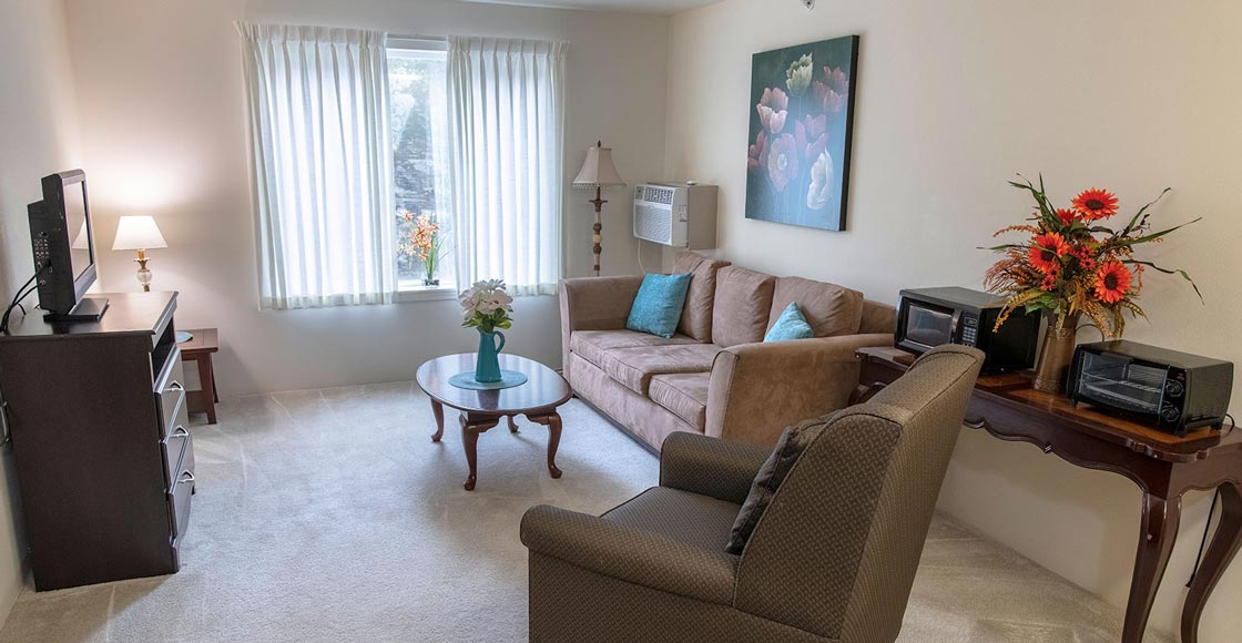 A well-lit living room with beige walls, featuring a large window with sheer curtains. The room has a brown sofa with blue cushions, a comfortable armchair, a wooden coffee table with a floral bouquet, a side table with a lamp, and a painting above the sofa.