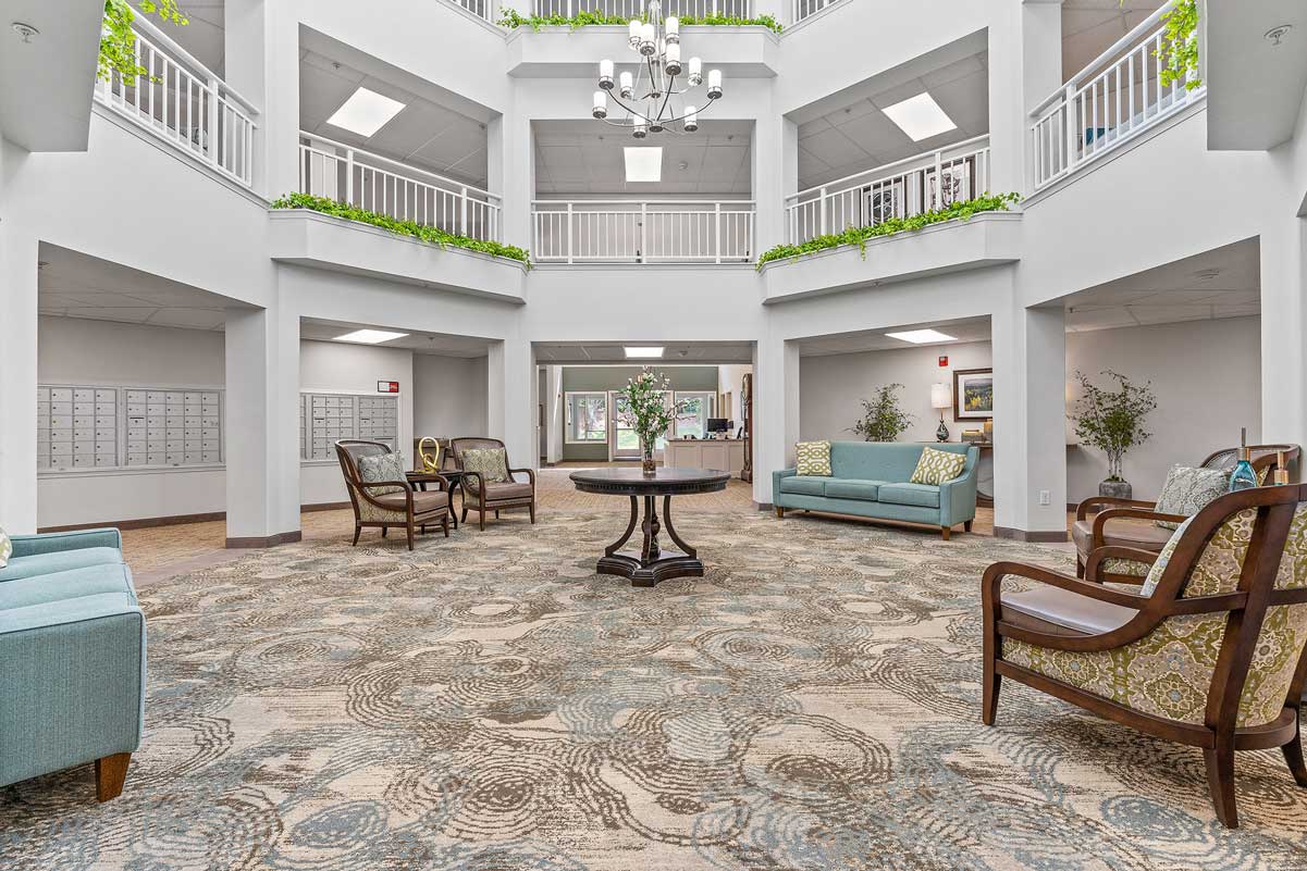 A spacious, well-lit lobby with a high ceiling and two floors of balconies featuring white railings. The area has several seating options, including sofas and armchairs. A central round table with flowers adds a decorative touch. Greenery adorns the balconies.