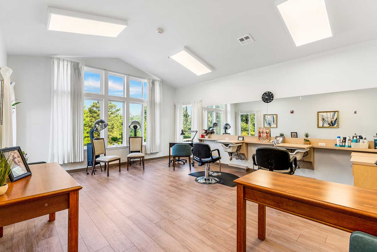 A bright and clean modern hair salon with large windows letting in ample natural light. The space features several workstations with chairs in front of mirrors, hair dryers, shelves with hair products, and wooden desks and chairs on a polished wood floor.