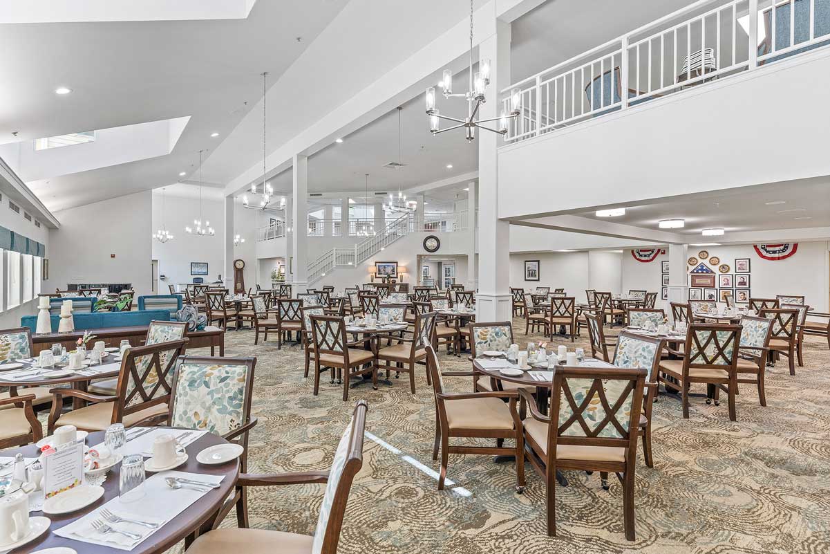 A spacious, well-lit dining area with numerous wooden tables and chairs arranged neatly. The room features high ceilings, two decorative chandeliers, and a mezzanine level with a white railing. Place settings and tableware are ready on the tables.