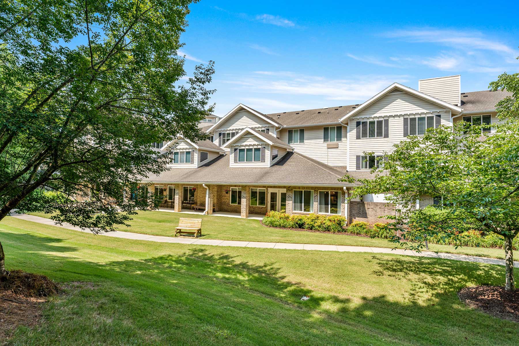 A large, two-story residential building with white siding and multiple windows. It is surrounded by green lawns, trees, and a few bushes. A wooden bench sits on a concrete pathway in the foreground. The sky is clear and blue.