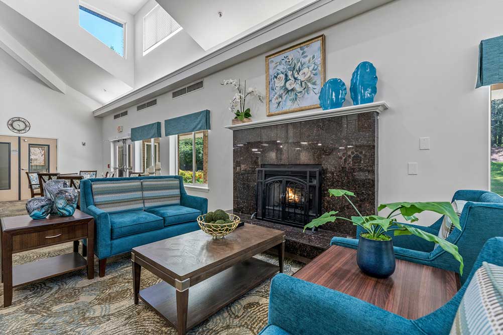 A cozy living room with skylights features a blue sofa set, patterned rug, wooden coffee and side tables, and a fireplace. Above the fireplace is a floral painting and blue vases. Large windows on the right bring in natural light and offer a view of greenery outside.