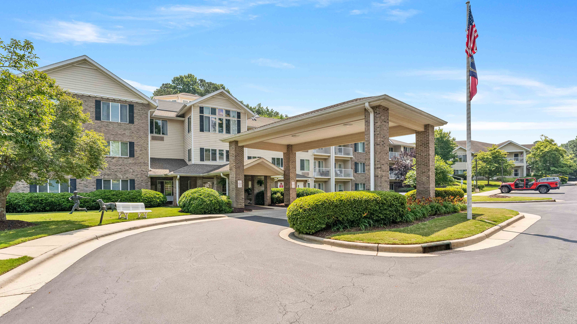 A three-story residential building with a covered entrance, surrounded by green shrubs and trees, a curved driveway, a bench, and an American flag on a tall flagpole. A red vehicle is parked nearby under a sunny sky.