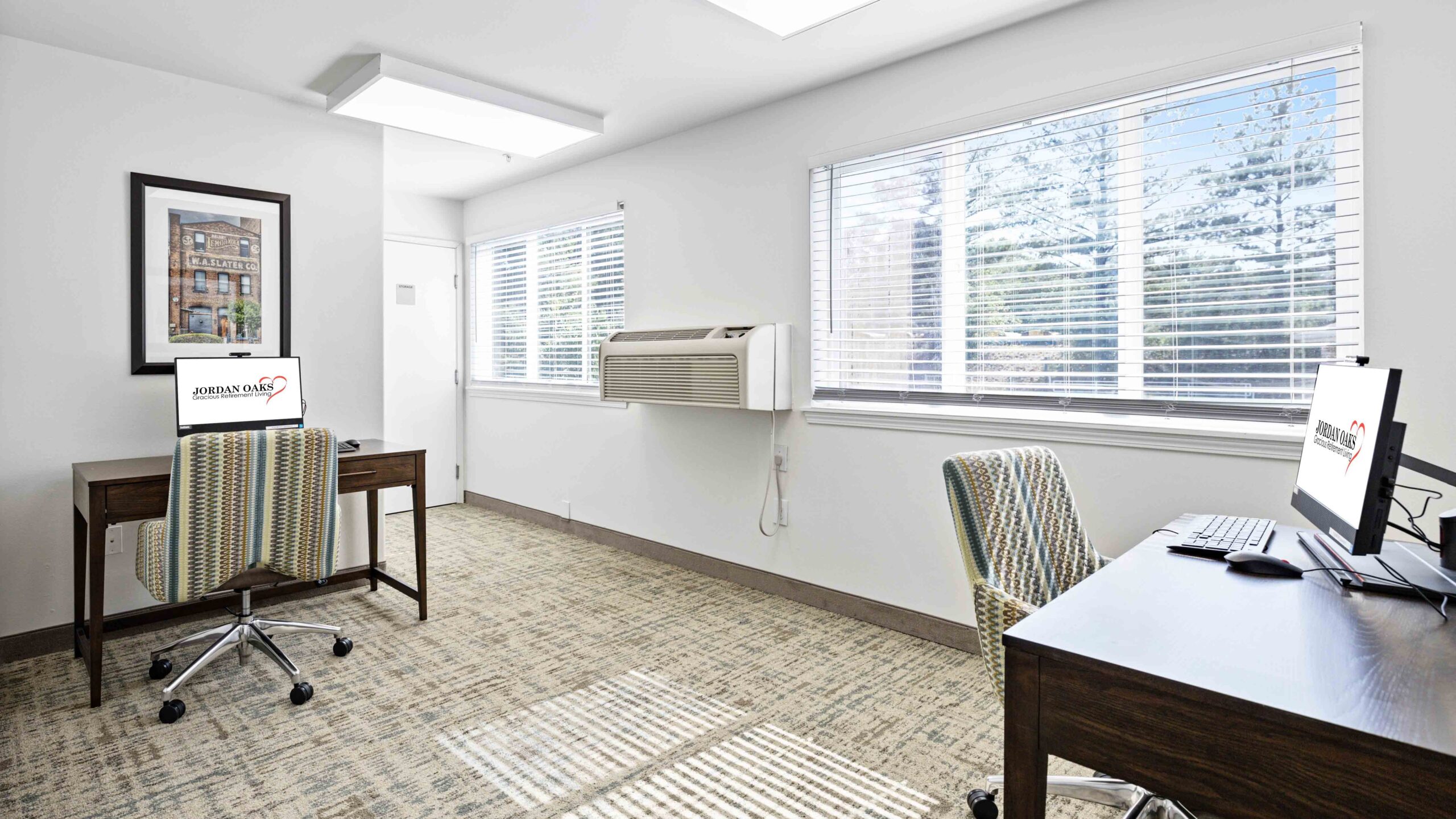 A bright office features two desks with computers and striped chairs, large windows letting in natural light, an air conditioning unit, and a framed picture on a white wall.