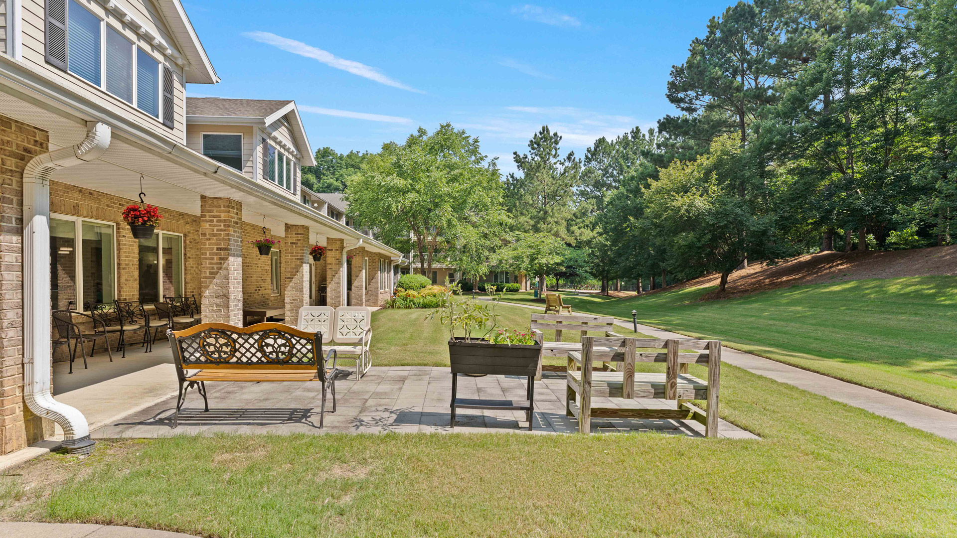 A sunny outdoor patio with benches and chairs sits beside a brick building, overlooking a grassy lawn and tree-lined walking path under a clear blue sky.