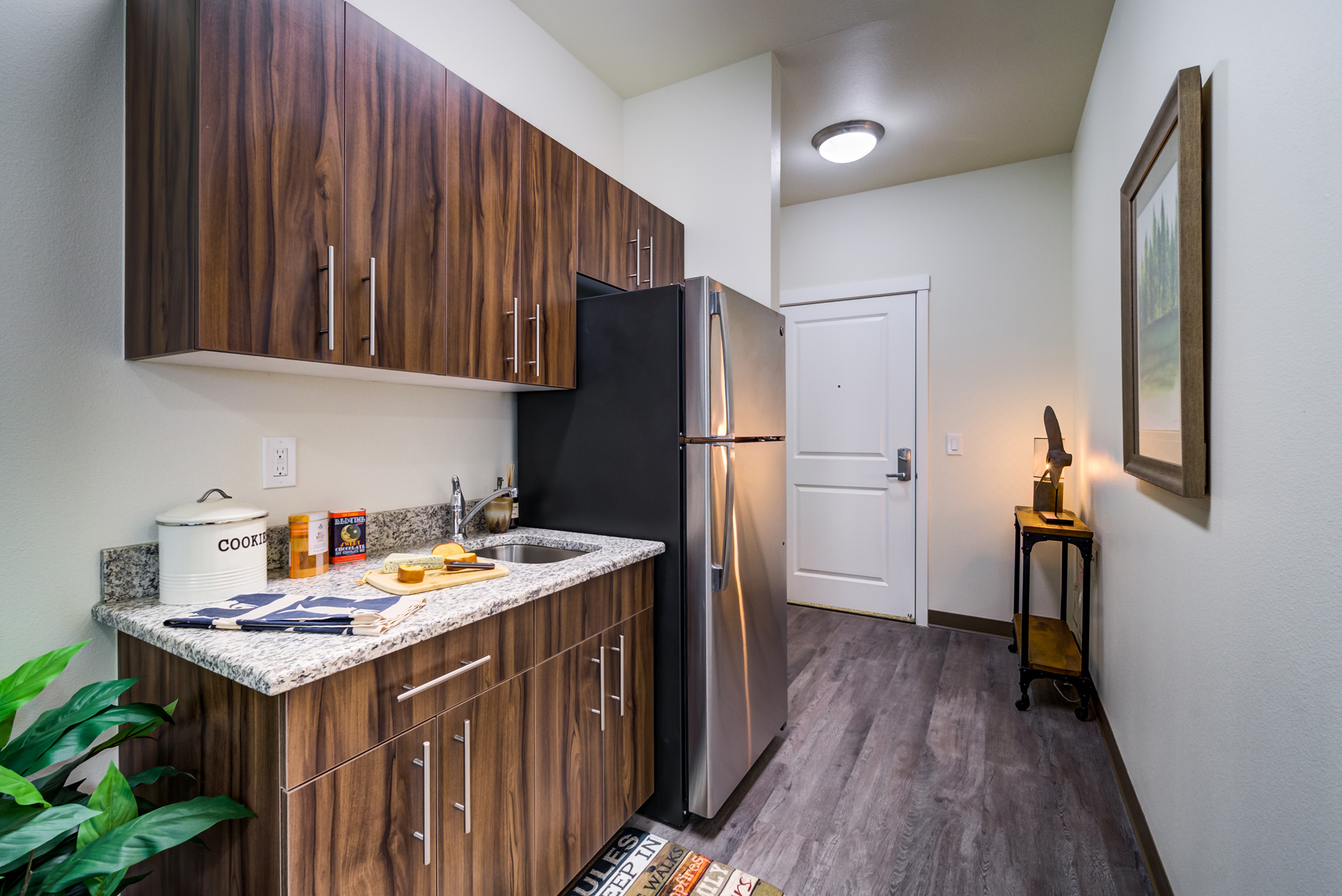 A modern, compact kitchen with wood cabinets, granite countertops, a stainless steel fridge, and a small sink. A cutting board with bread, a “COOKIES” jar, and kitchen decor sit on the counter. The entry door is visible in the background.