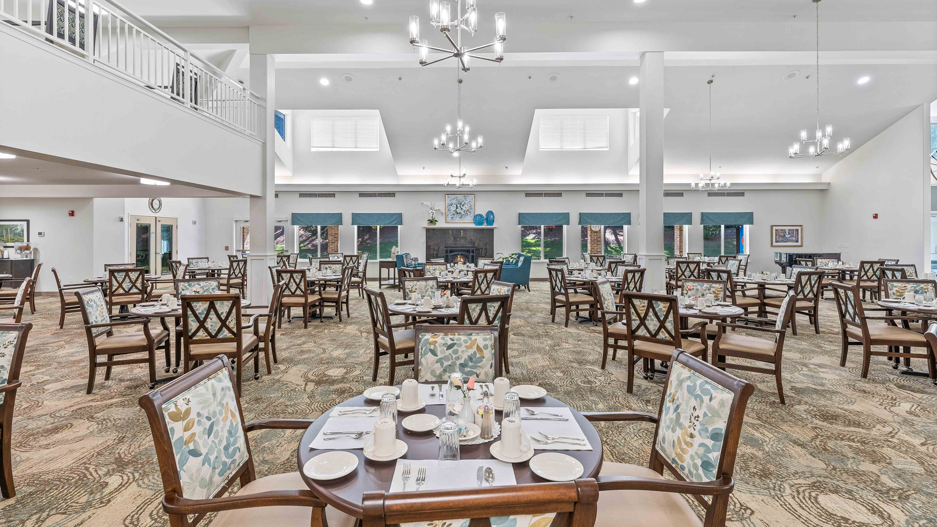 Spacious, well-lit dining hall with neatly arranged wooden tables and padded chairs, set for a meal; chandeliers hang from the high ceiling and large windows line the back wall.