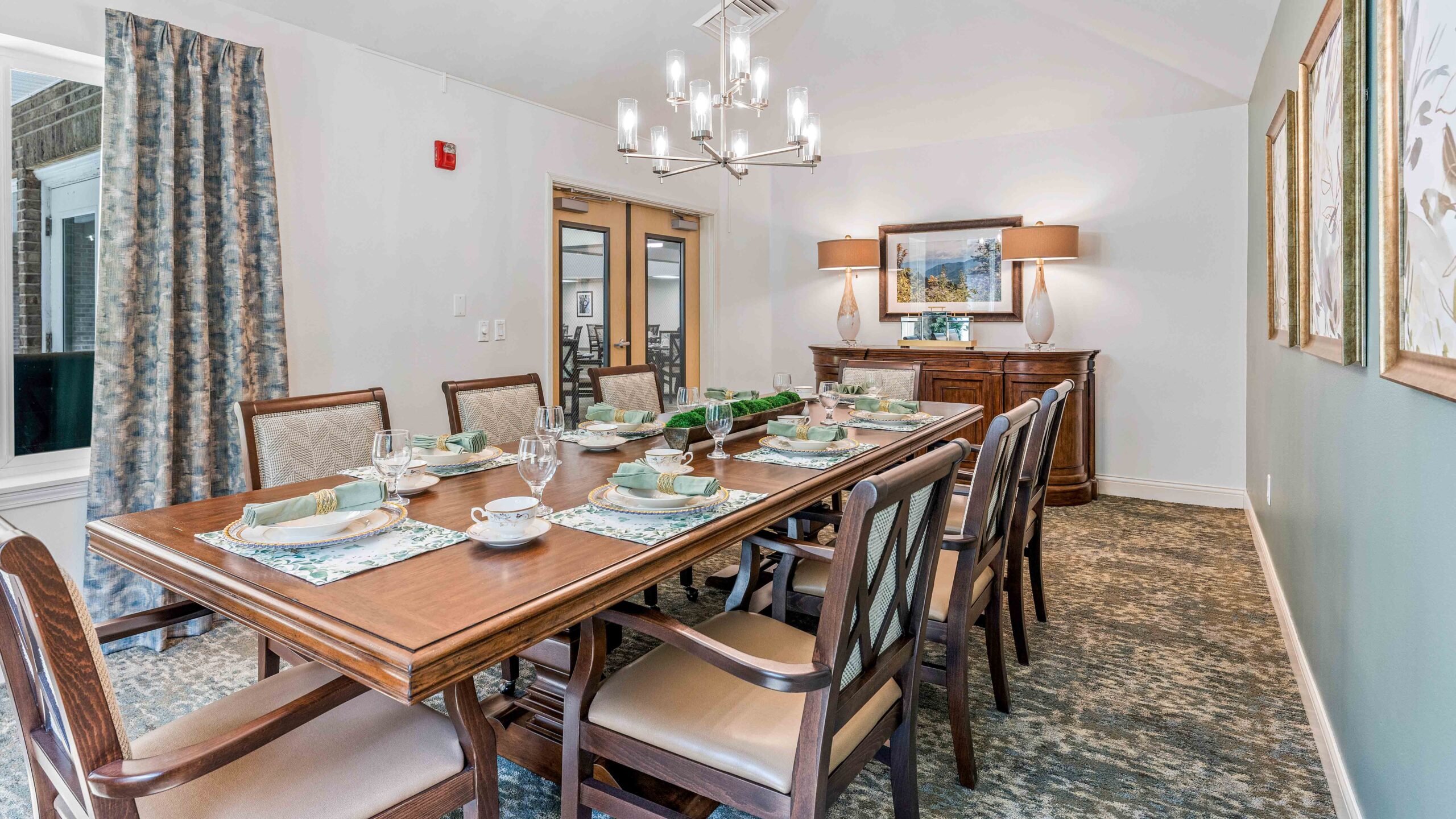 A formal dining room with a long wooden table set for eight, elegant chairs, a chandelier, artwork on the walls, a sideboard with lamps, and large windows with drapes.