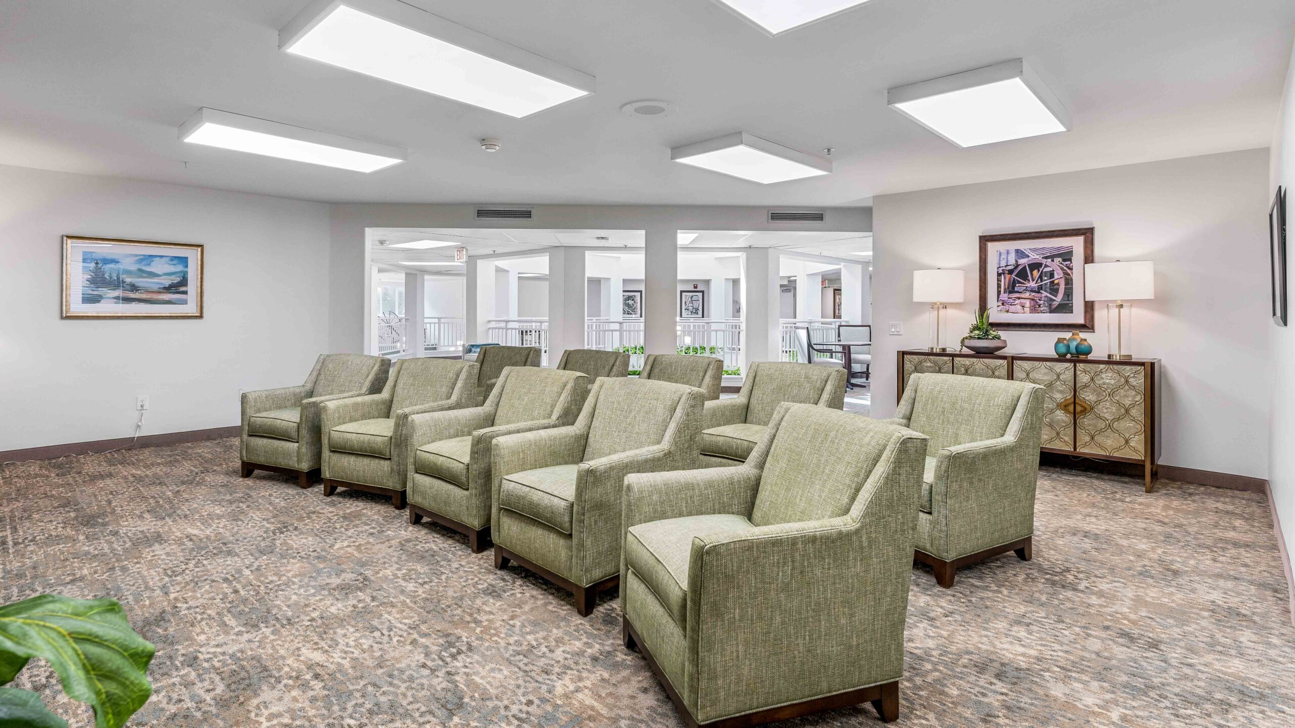 A bright, modern waiting area with eight green upholstered armchairs arranged in two rows, neutral carpet, artwork on white walls, and large ceiling lights. A console table with lamps and decor sits against the back wall.