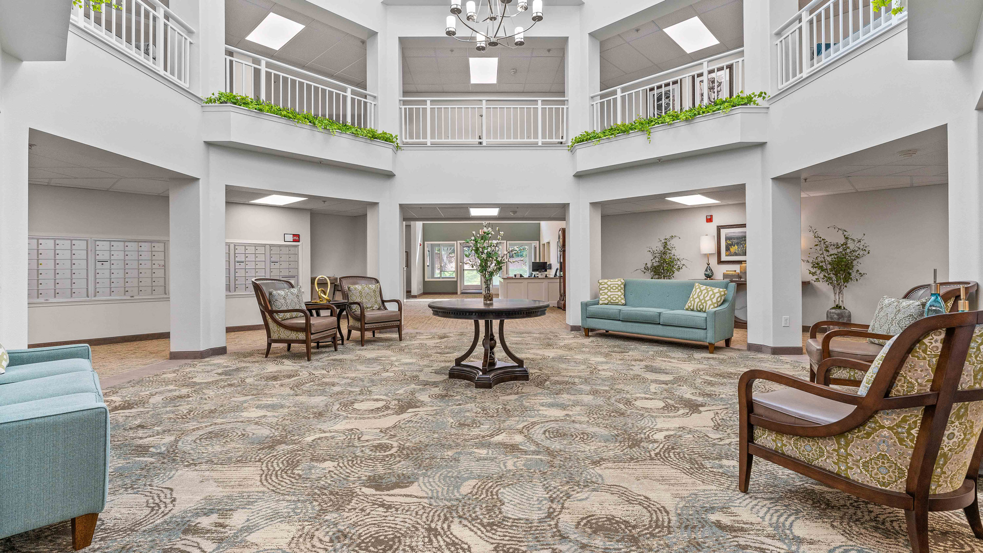 Bright, spacious lobby with patterned carpet, comfortable armchairs, teal sofas, a round table with a flower vase, and balconies on the upper level. White walls, green plants, and natural light create a welcoming atmosphere.