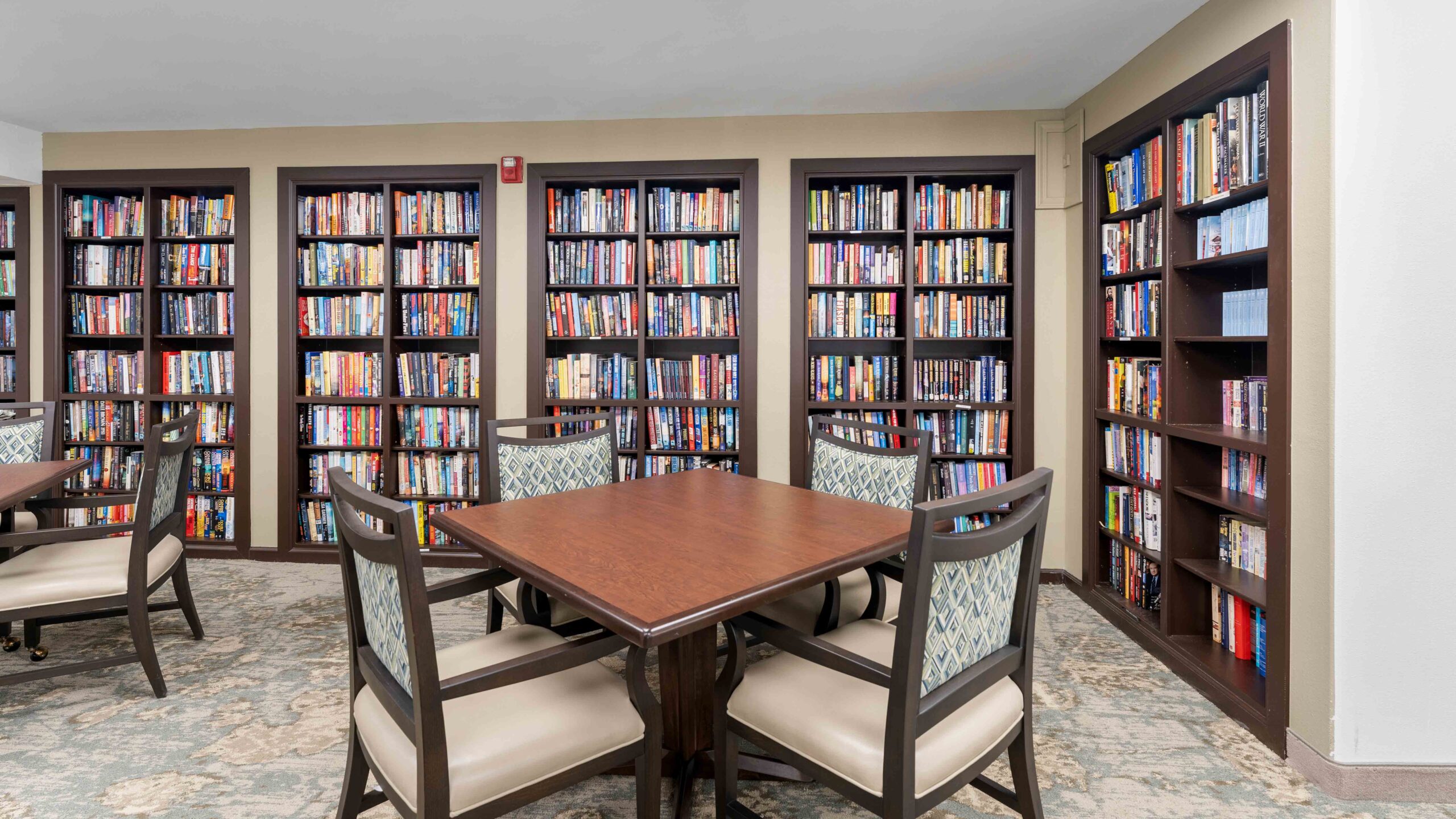 A quiet library room with four wooden chairs around a square table, and walls lined with tall bookshelves filled with colorful books. The carpeted floor and soft lighting create a cozy atmosphere.
