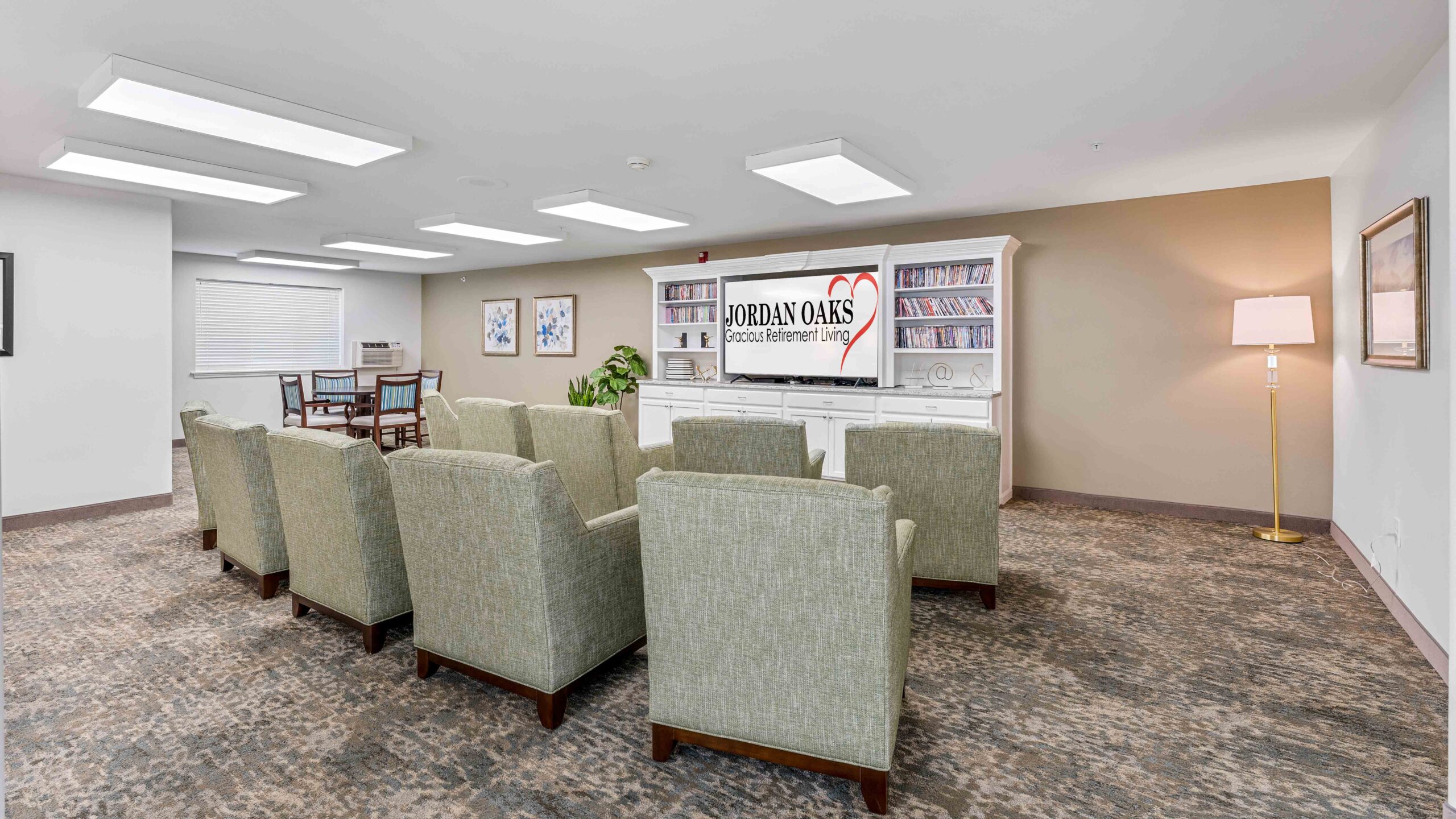 A small, neatly arranged room with six green armchairs facing a white entertainment center with a TV showing the "Jordan Oaks Retirement Living" logo. Bookshelves, a table with chairs, wall art, and a floor lamp decorate the space.