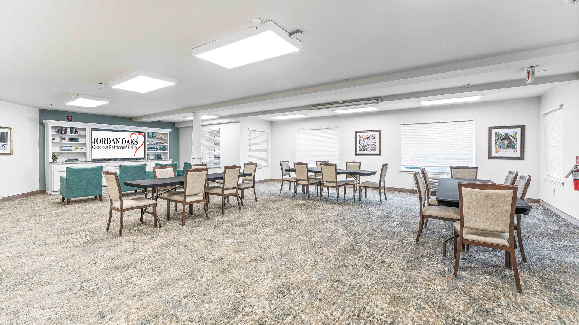 A spacious, well-lit community room with multiple tables and chairs, teal armchairs near shelves, framed art on the walls, and a sign reading "Jordan Oaks" above a white cabinet.