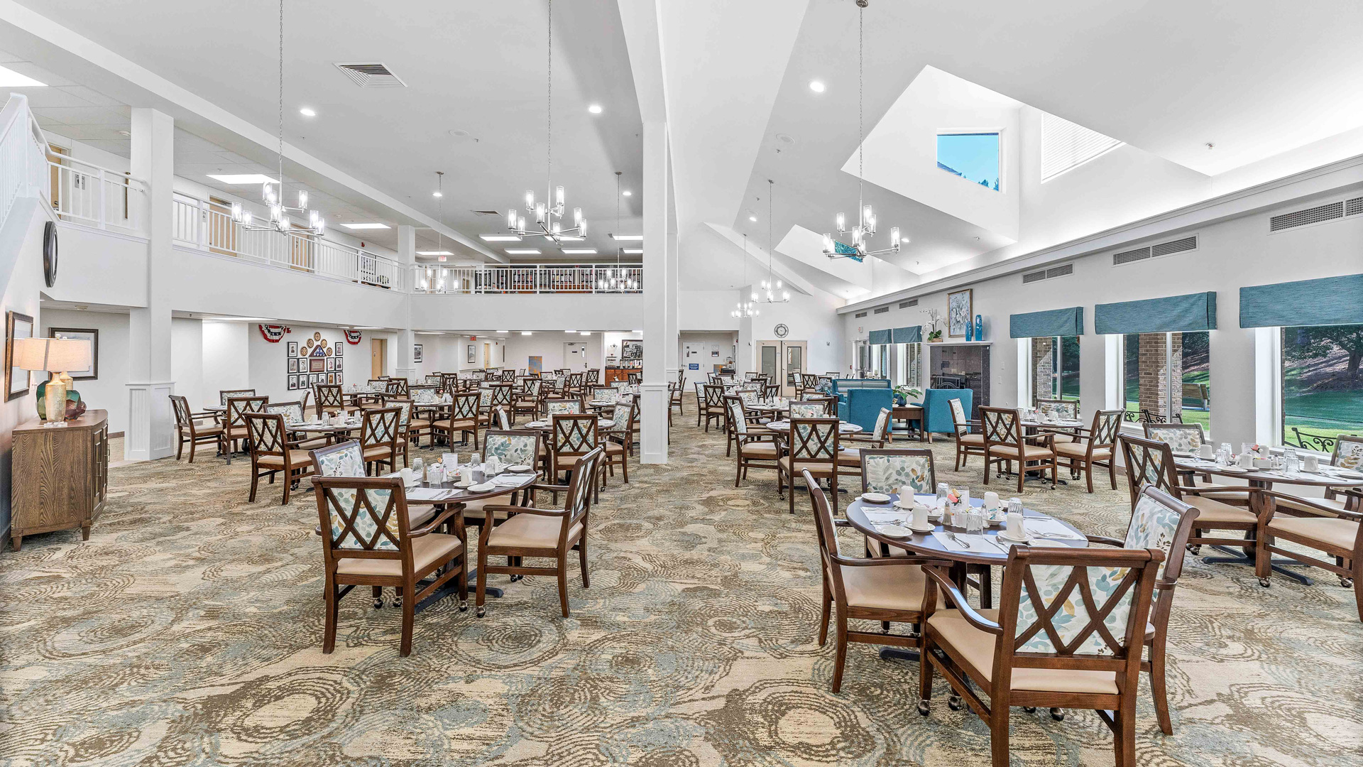 A spacious, well-lit dining room with many neatly arranged tables and chairs, elegant chandeliers, large windows, and high ceilings. Place settings are ready for guests, and blue accents add to the inviting atmosphere.