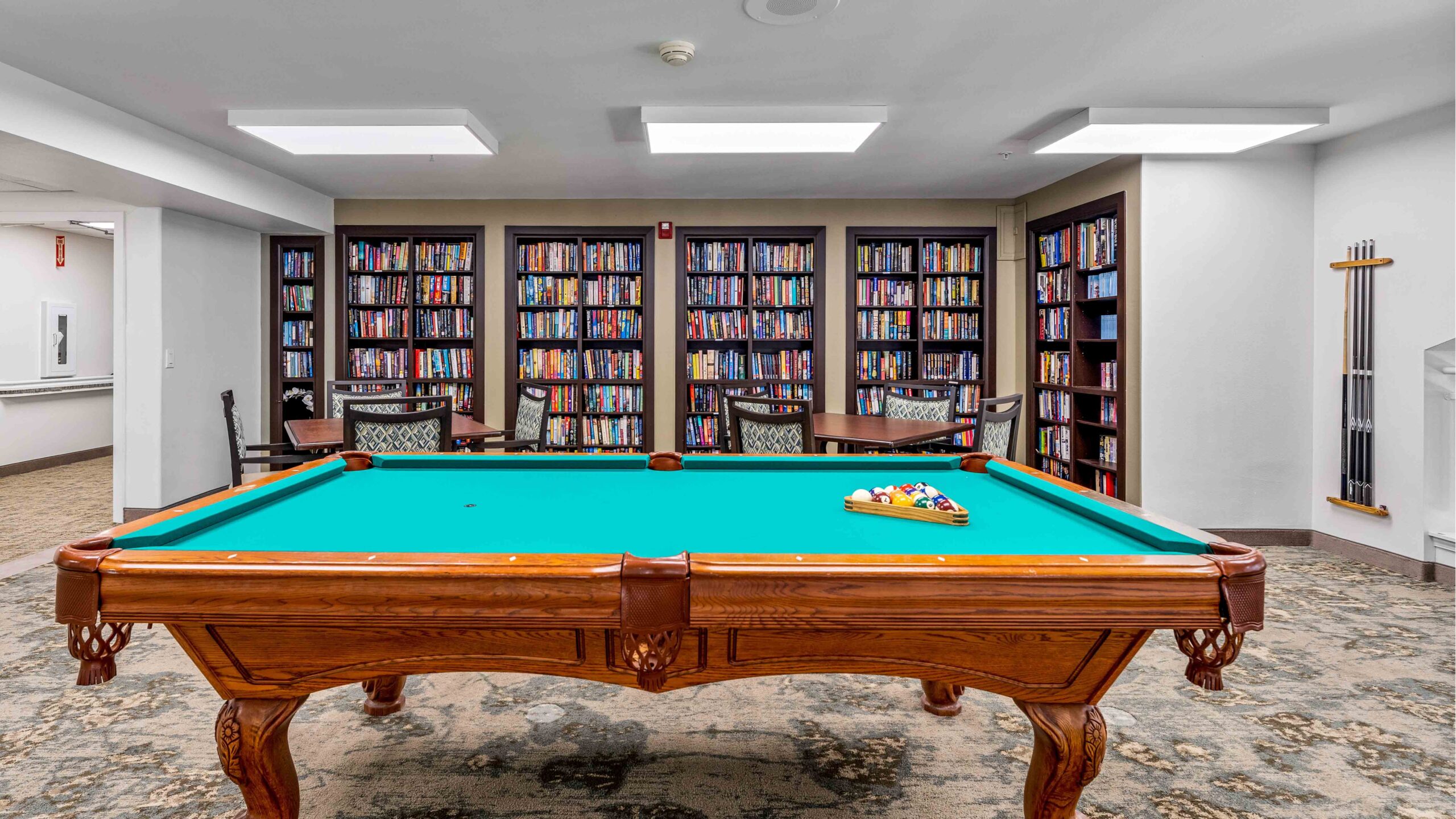 A pool table with balls racked and cues on the wall is centered in a room with four chairs and bookshelves filled with books in the background. The space is well-lit with modern ceiling lights.