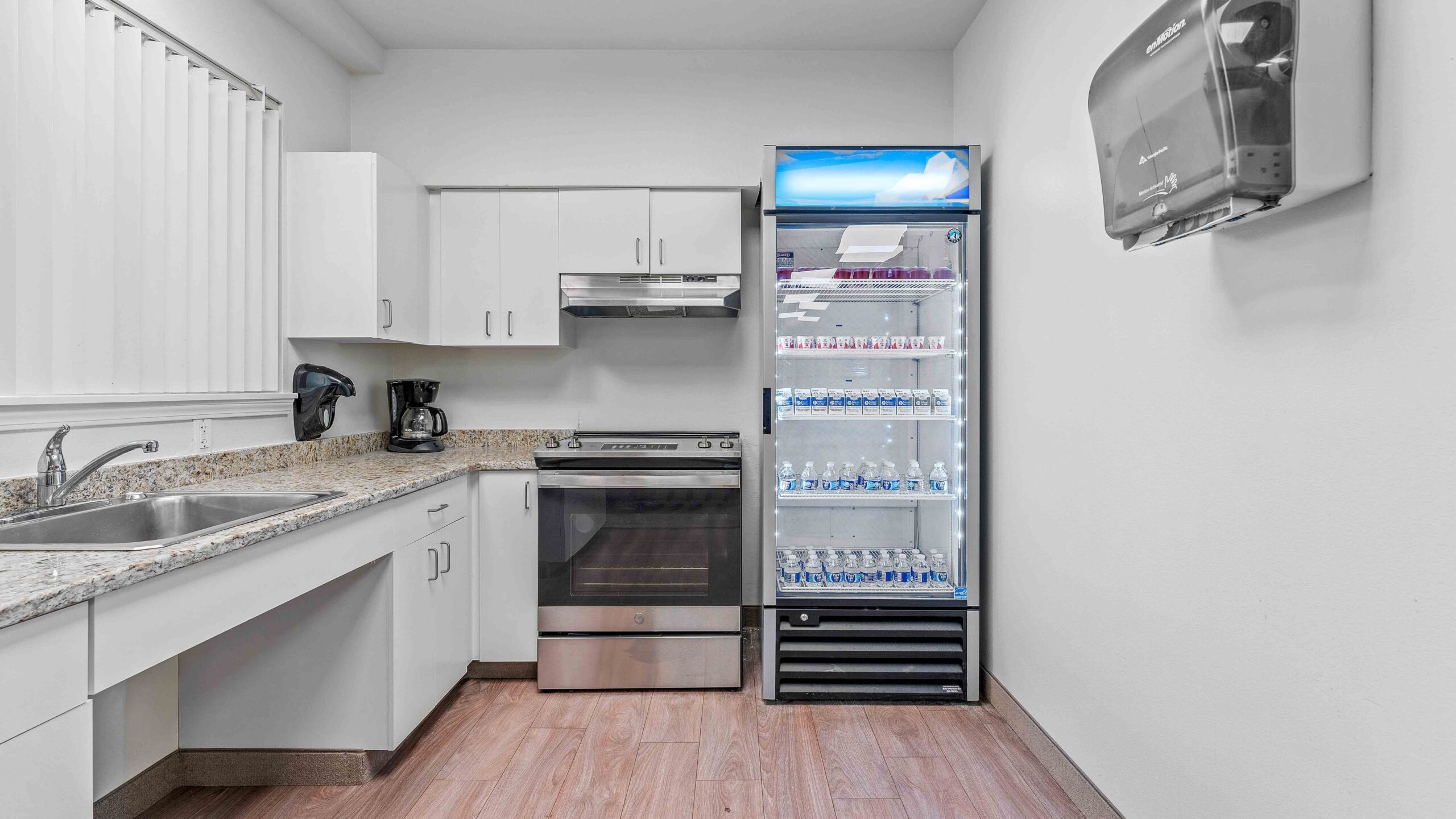 A clean, modern kitchenette with white cabinets, granite countertops, an oven, a coffee maker, and a glass-front refrigerator stocked with bottled water. A paper towel dispenser is mounted on the wall.