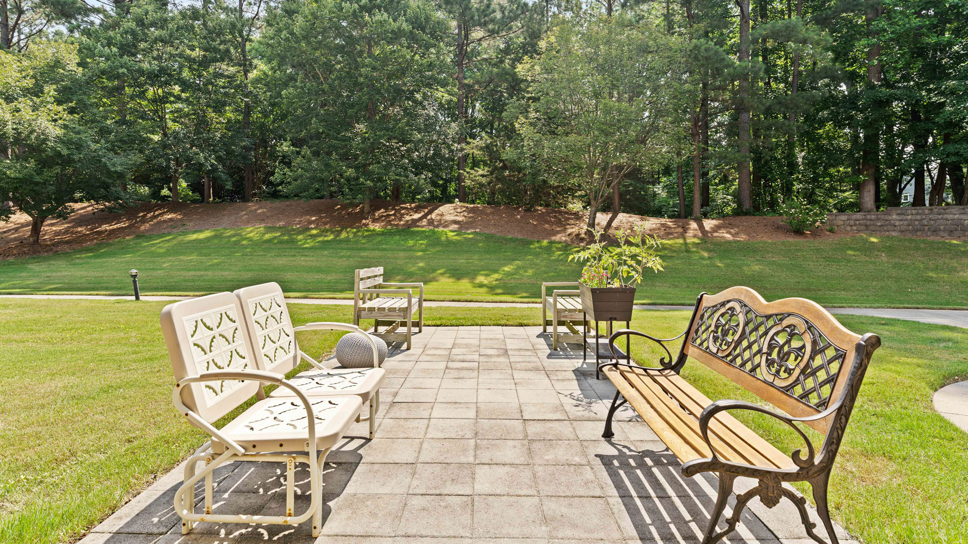 A sunny outdoor patio with metal chairs, a wooden bench, and a potted plant on a paved area surrounded by green grass and trees in the background.