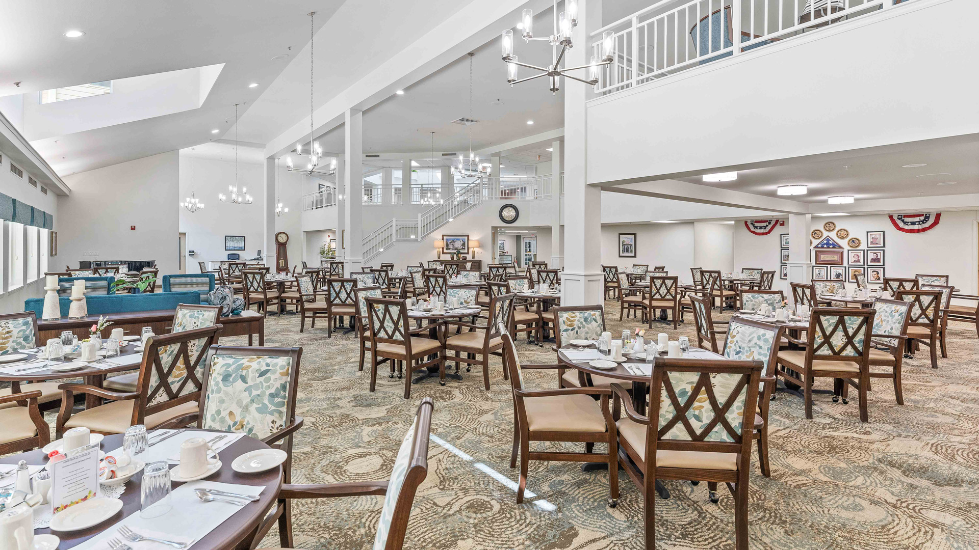 Bright, spacious dining hall with neatly arranged wooden tables and chairs, set for a meal. The room has chandeliers, framed pictures on the walls, and a staircase leading to an upper level with railings.