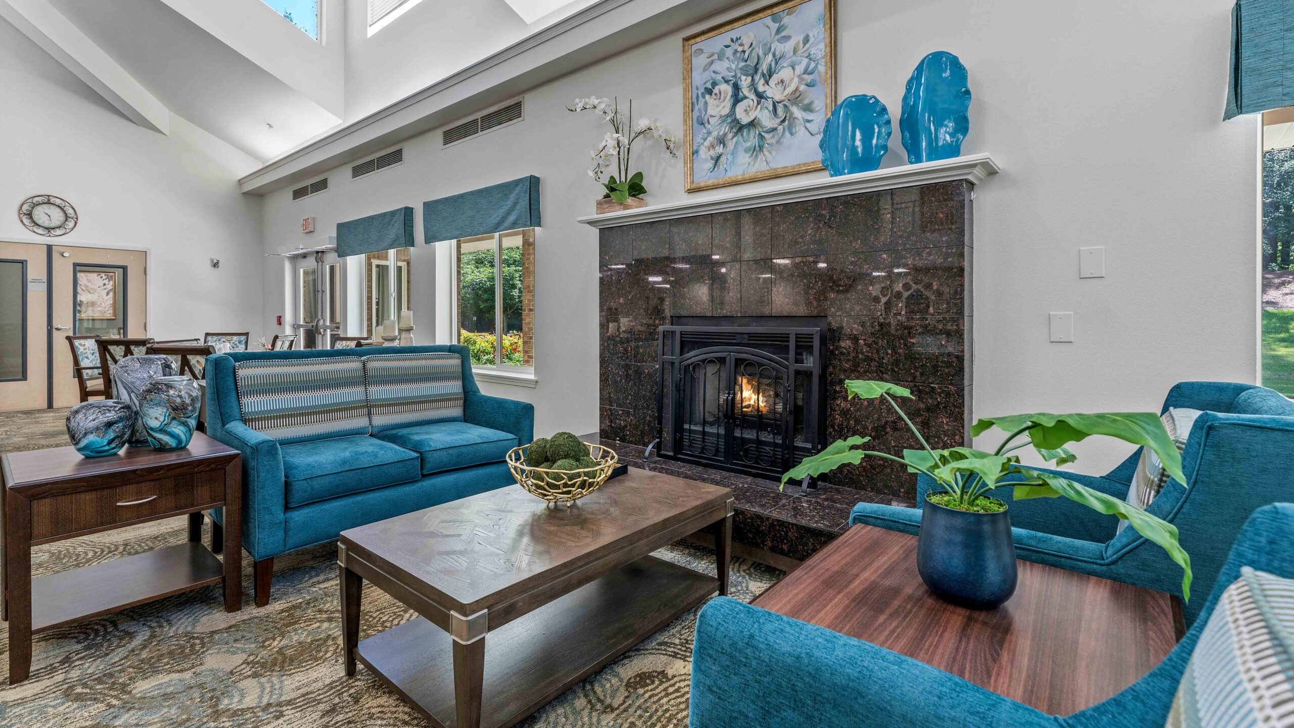 A cozy living room with blue sofas, a dark marble fireplace, a coffee table, houseplants, and large windows letting in natural light. Blue vases and floral artwork decorate the mantel above the fireplace.