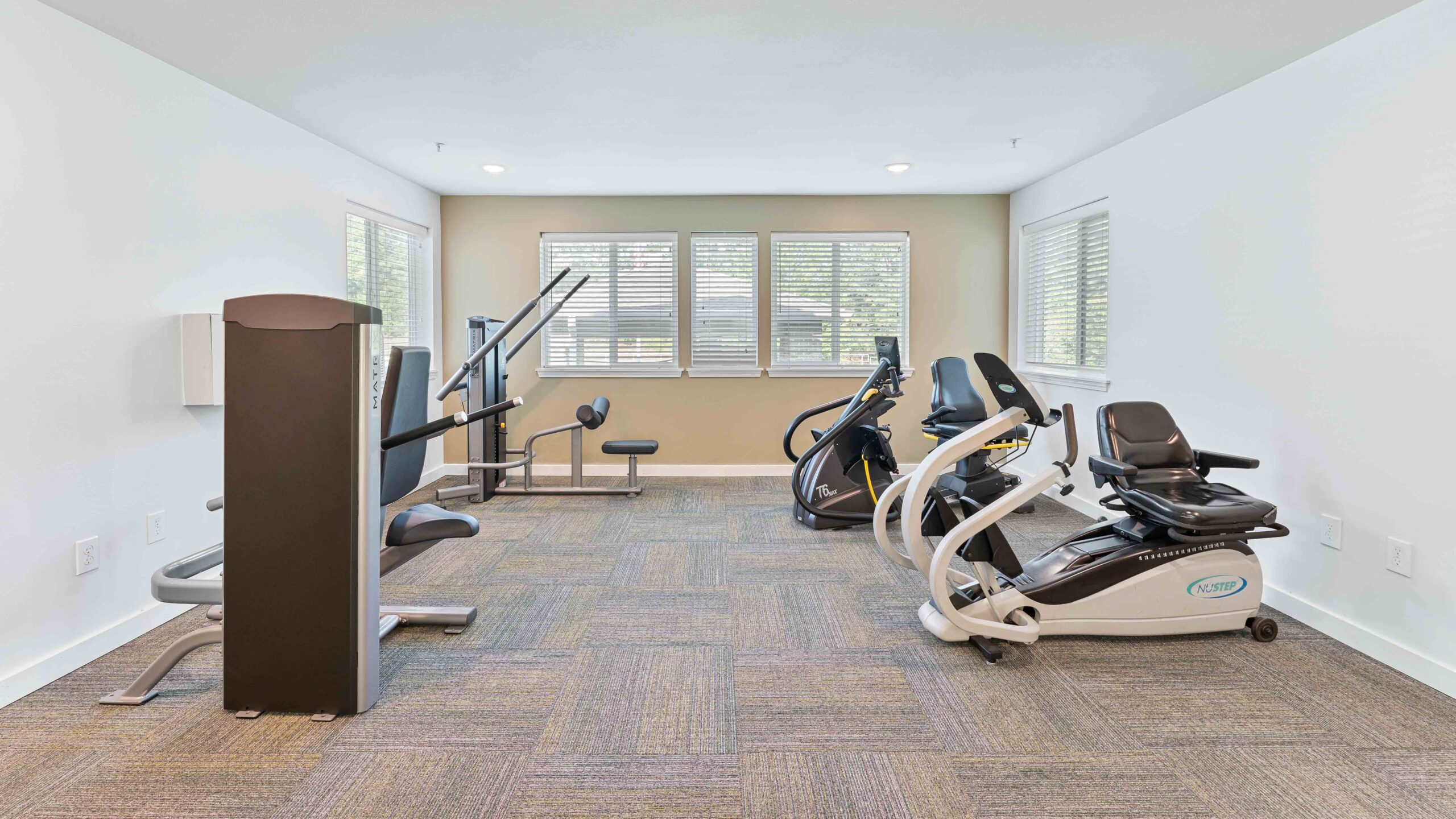 A bright, clean fitness room with large windows, two recumbent exercise bikes, a stationary bike, a weight machine, and a bench on carpeted flooring.