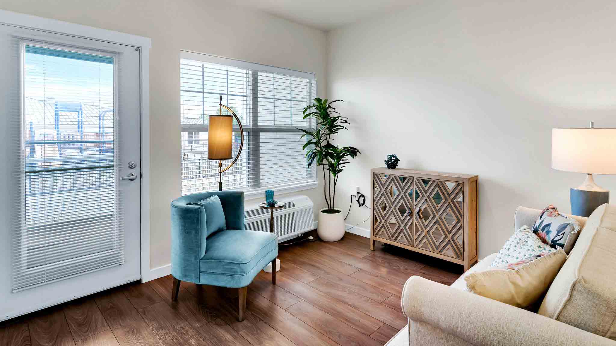 A bright living room with wood floors, a beige sofa, a blue accent chair, a modern lamp, a patterned sideboard, a potted plant, and large windows with blinds letting in natural light.