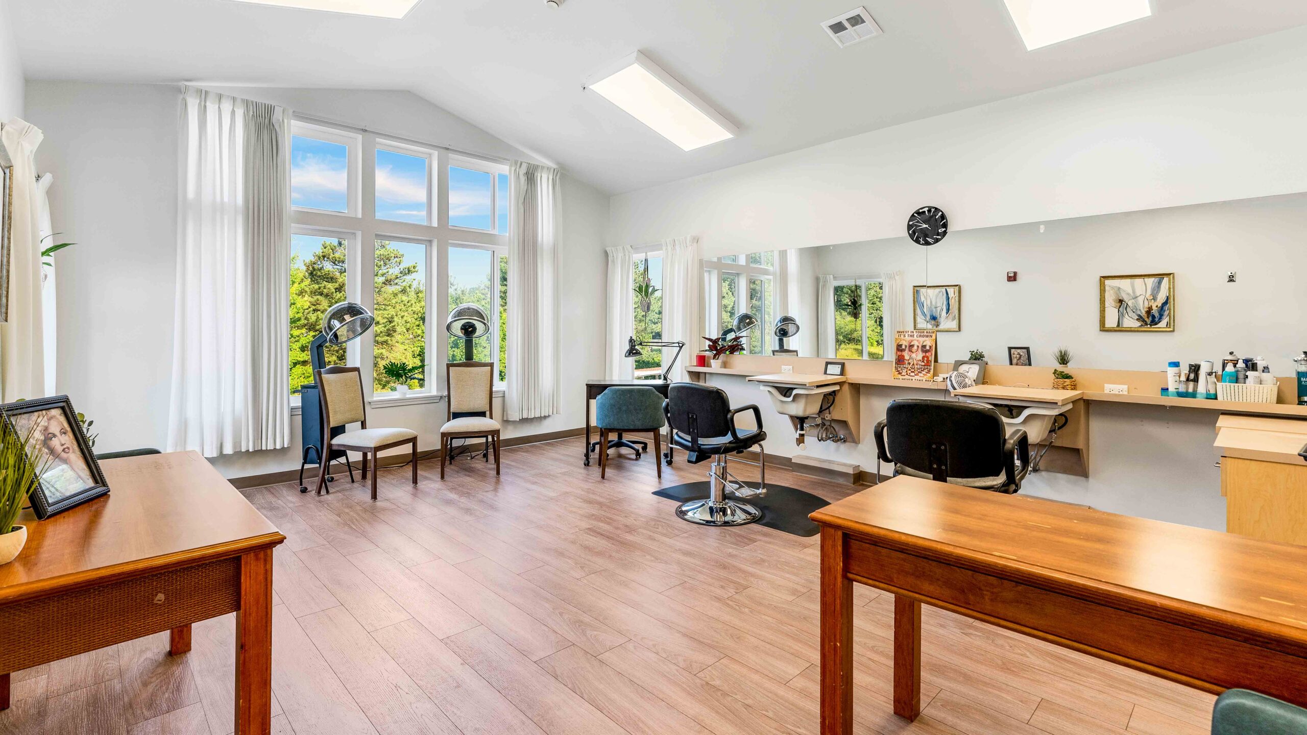 Bright, modern salon with large windows, wooden floors, several chairs, mirrors, and tables. Natural light fills the room, which is decorated with plants and framed pictures, creating a welcoming and tidy atmosphere.