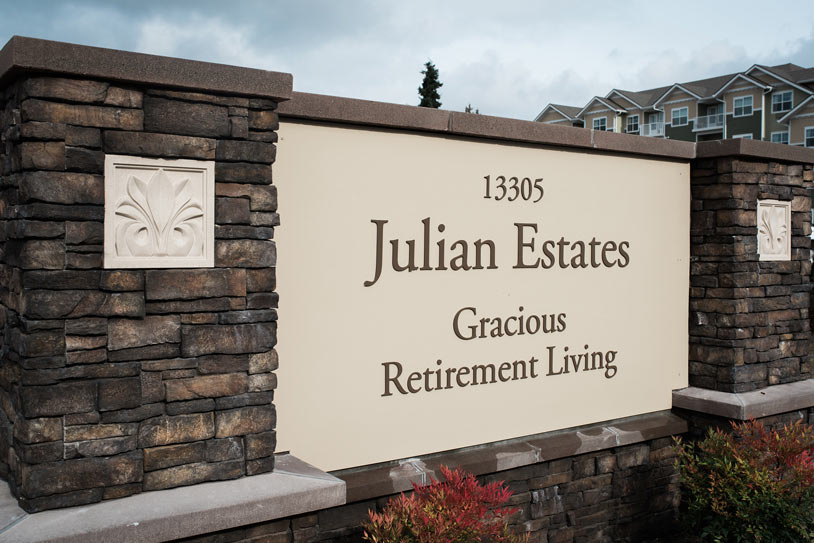 A stone monument sign with the text "13305 Julian Estates Gracious Retirement Living" inscribed on a beige backdrop. The sign is framed by two stone pillars, each adorned with a decorative square panel. Townhouses are visible in the background against a cloudy sky.
