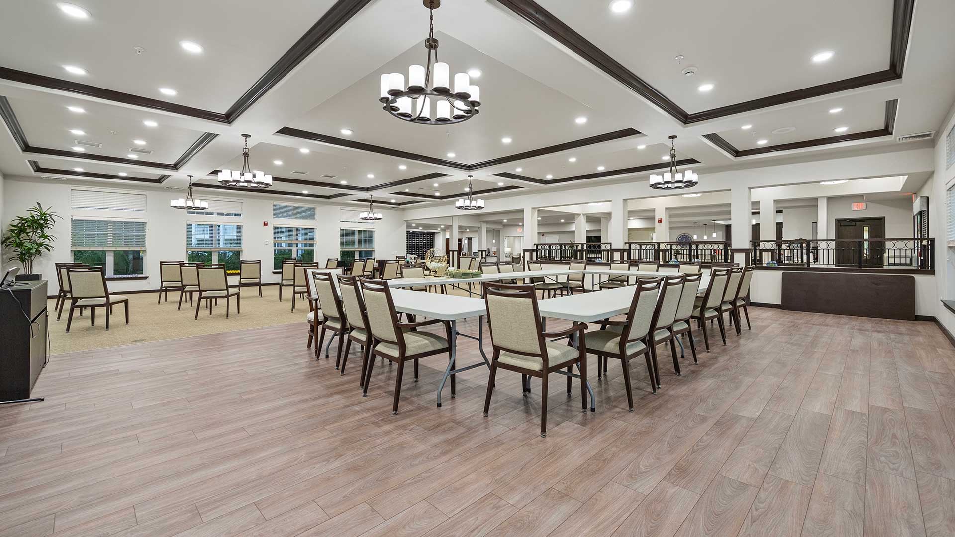 A spacious, modern dining room with a tiled floor and several rows of tables and chairs. Elegant chandeliers hang from the ceiling. Large windows allow natural light to enter the room.