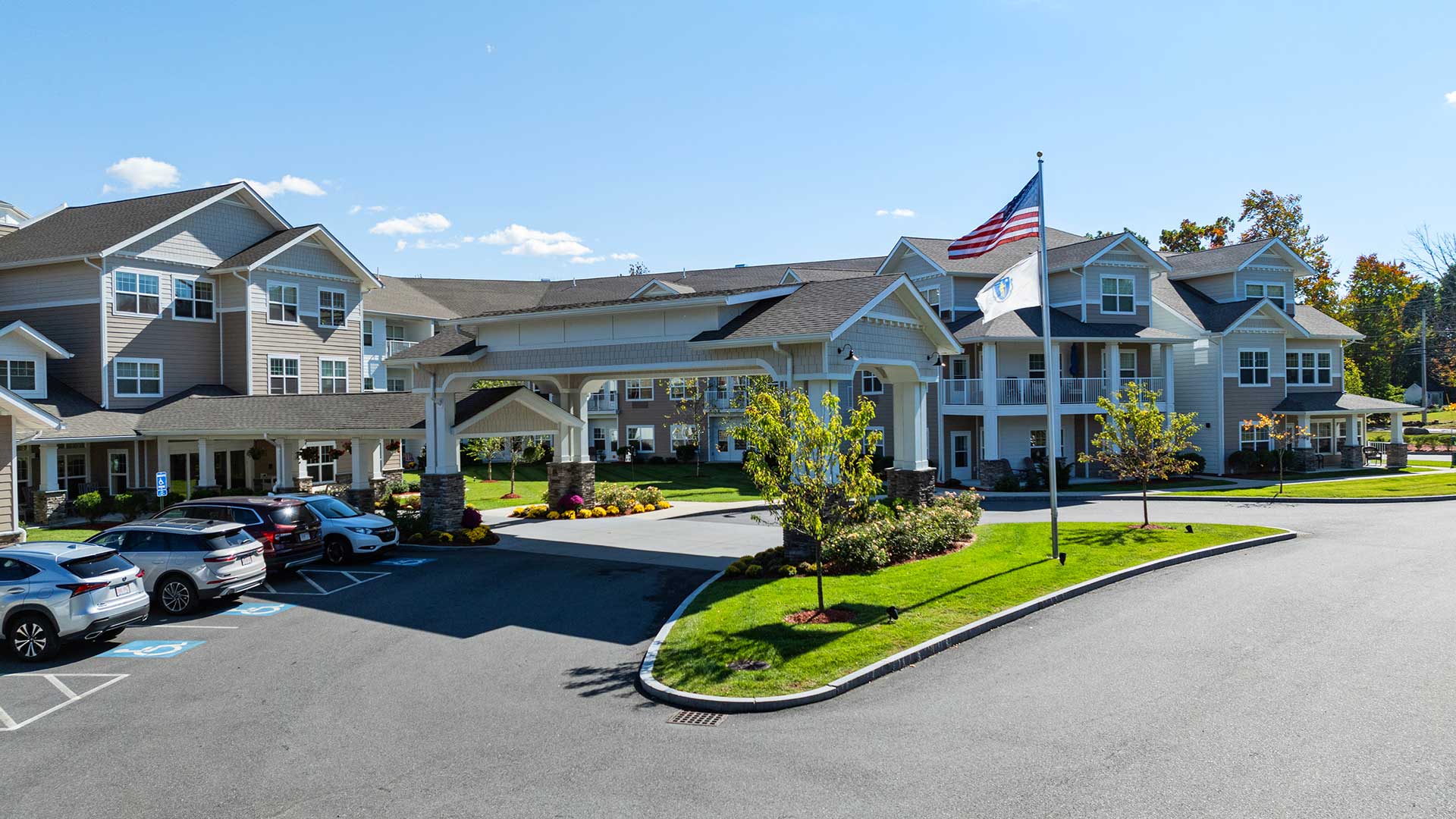 A large, multi-story assisted living facility with a grand entrance and covered driveway. An American flag is prominently displayed in front. Several cars are parked nearby, and trees with autumn foliage enhance the scene under a clear blue sky.