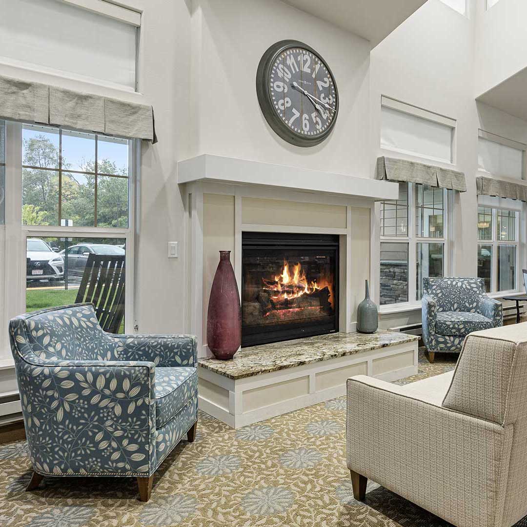 A cozy living room featuring a lit fireplace with a large decorative clock above it. Two patterned blue armchairs and a beige sofa surround the fireplace. Large windows with gray valances provide natural light, and a patterned rug covers the floor.