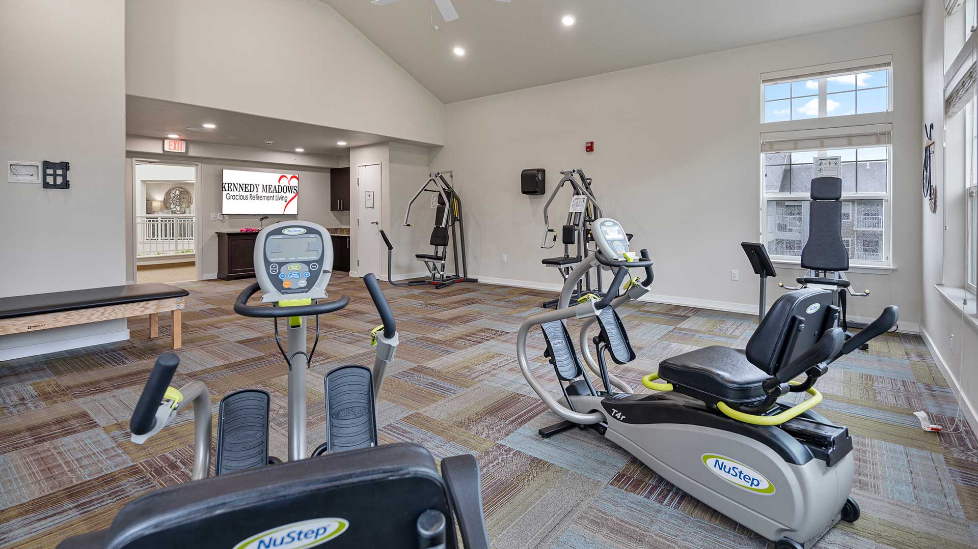 A fitness room with exercise equipment including NuStep machines and a weight lifting area. The room has large windows, carpeted floors, and neutral-colored walls. A sign in the background reads 