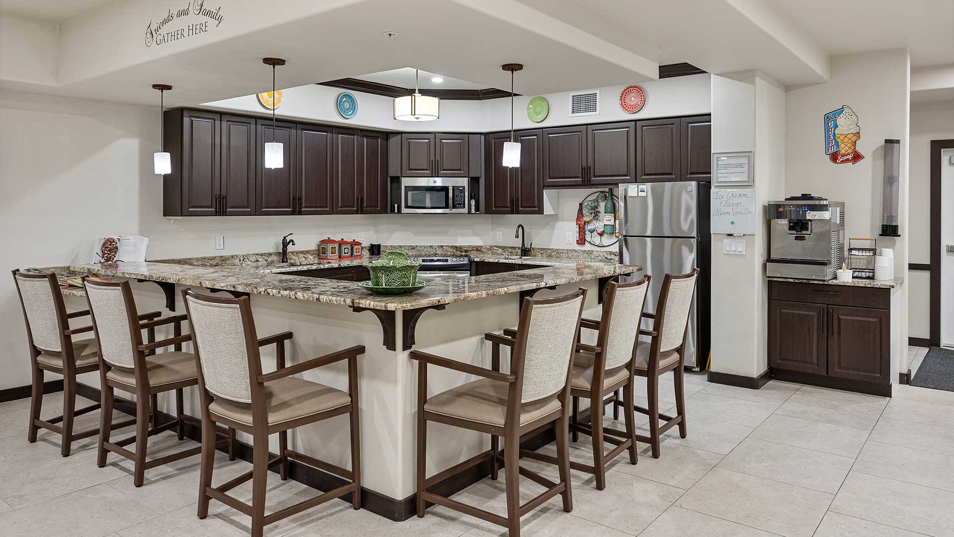 A modern kitchen with dark wood cabinets and granite countertops. The kitchen features a central island with bar seating for six, stainless steel appliances, and pendant lighting. A coffee machine is on a side counter next to a doorway.