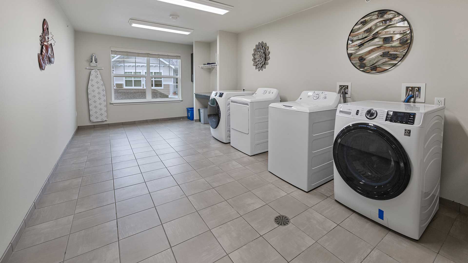 A laundry room with three washing machines lined up against a beige wall. An ironing board and iron are on the left, while circular wall art decorates the room. A large window at the end of the room lets in natural light. The floor is tiled.