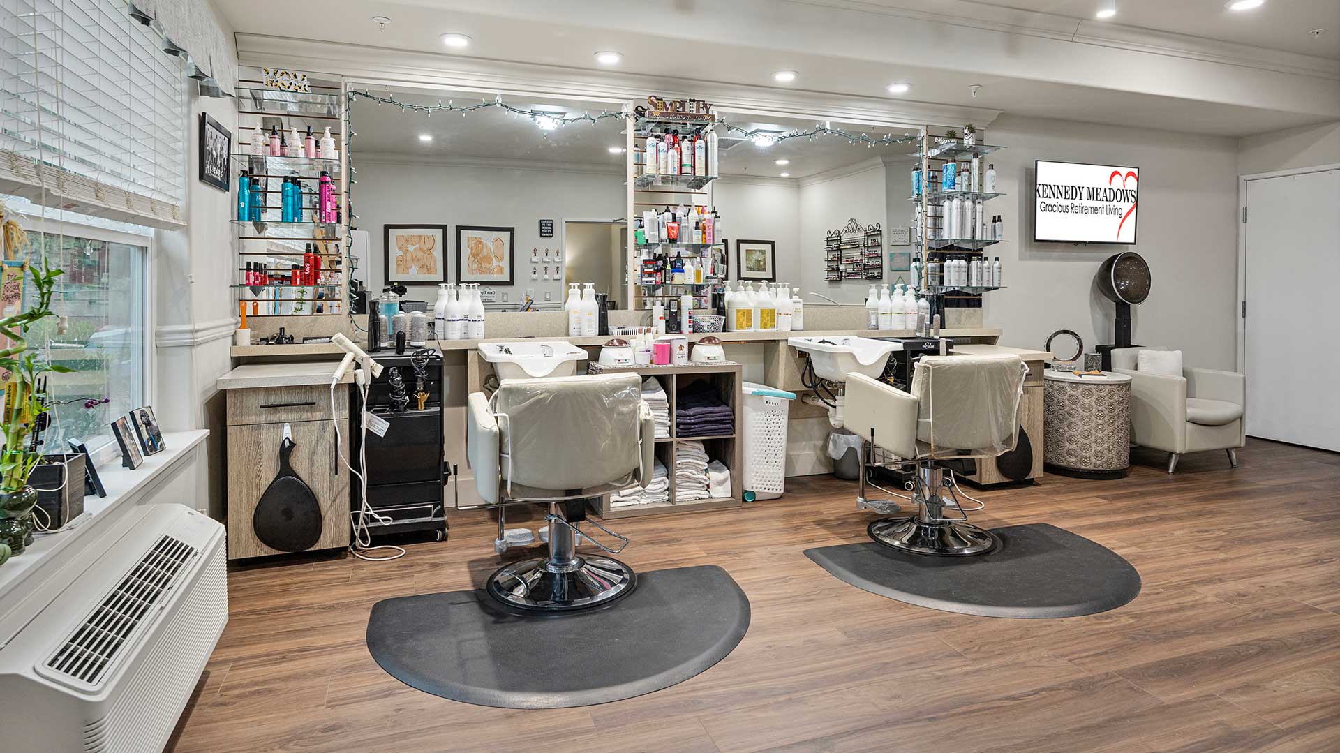 A modern hair salon interior featuring two styling chairs in front of large mirrors. Shelves are stocked with various hair products, and the walls are decorated with artwork. The space is well-lit and has a polished, wooden floor.